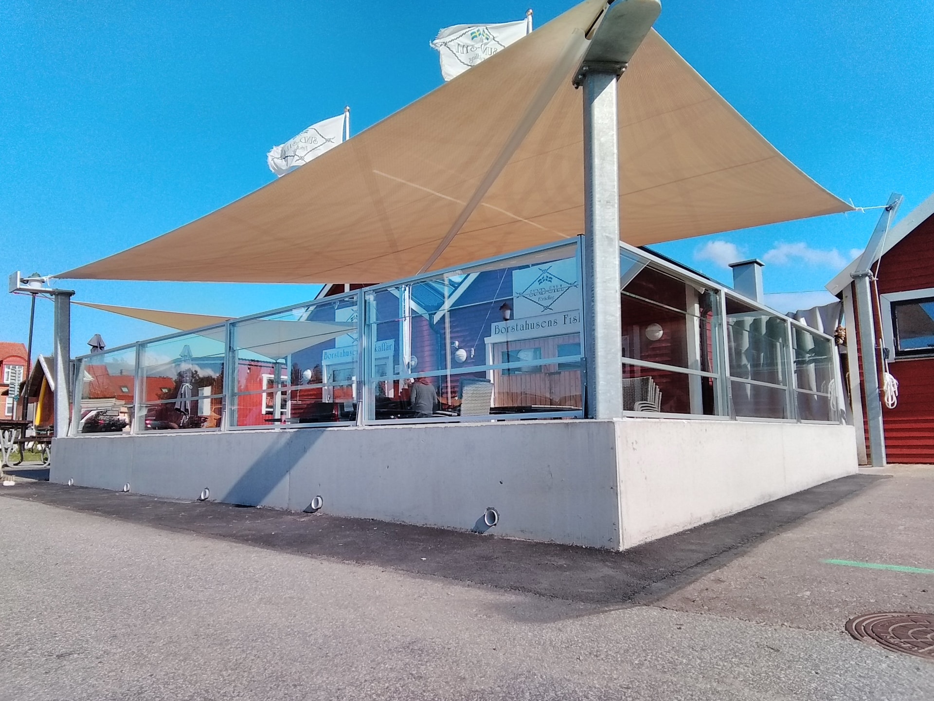 Beigt triangelformat solsegel vid kustrestaurang med glasräcke och röda trähus i bakgrunden