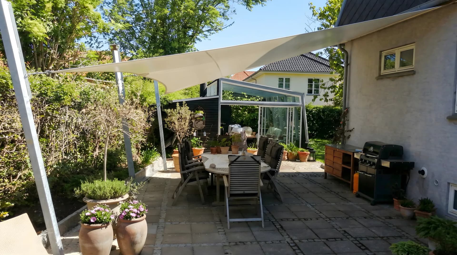 Vitt triangelformat solsegel på stenlagd gård med trädgårdsstolar, grill och krukväxter