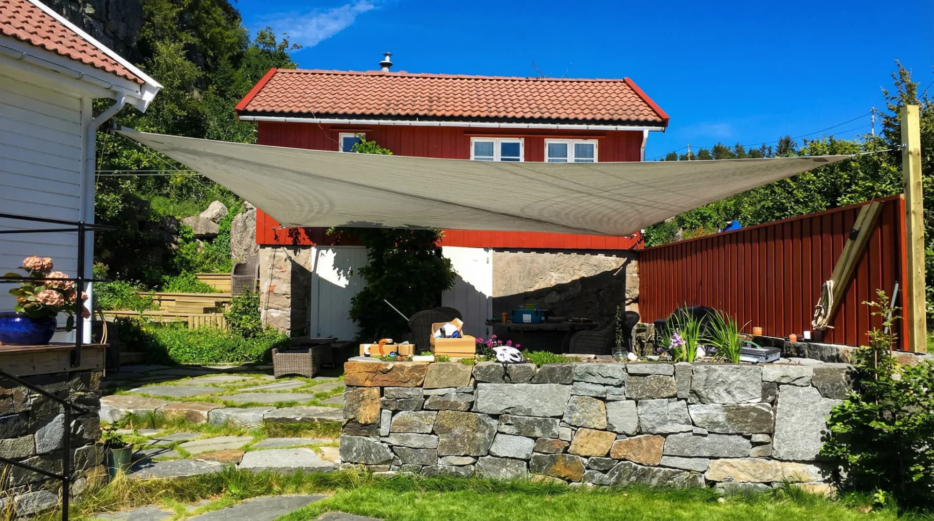 Beigt solsegel spännt på uteservering vid glasskärm och röda trähus vid vattnet i kvällsljus