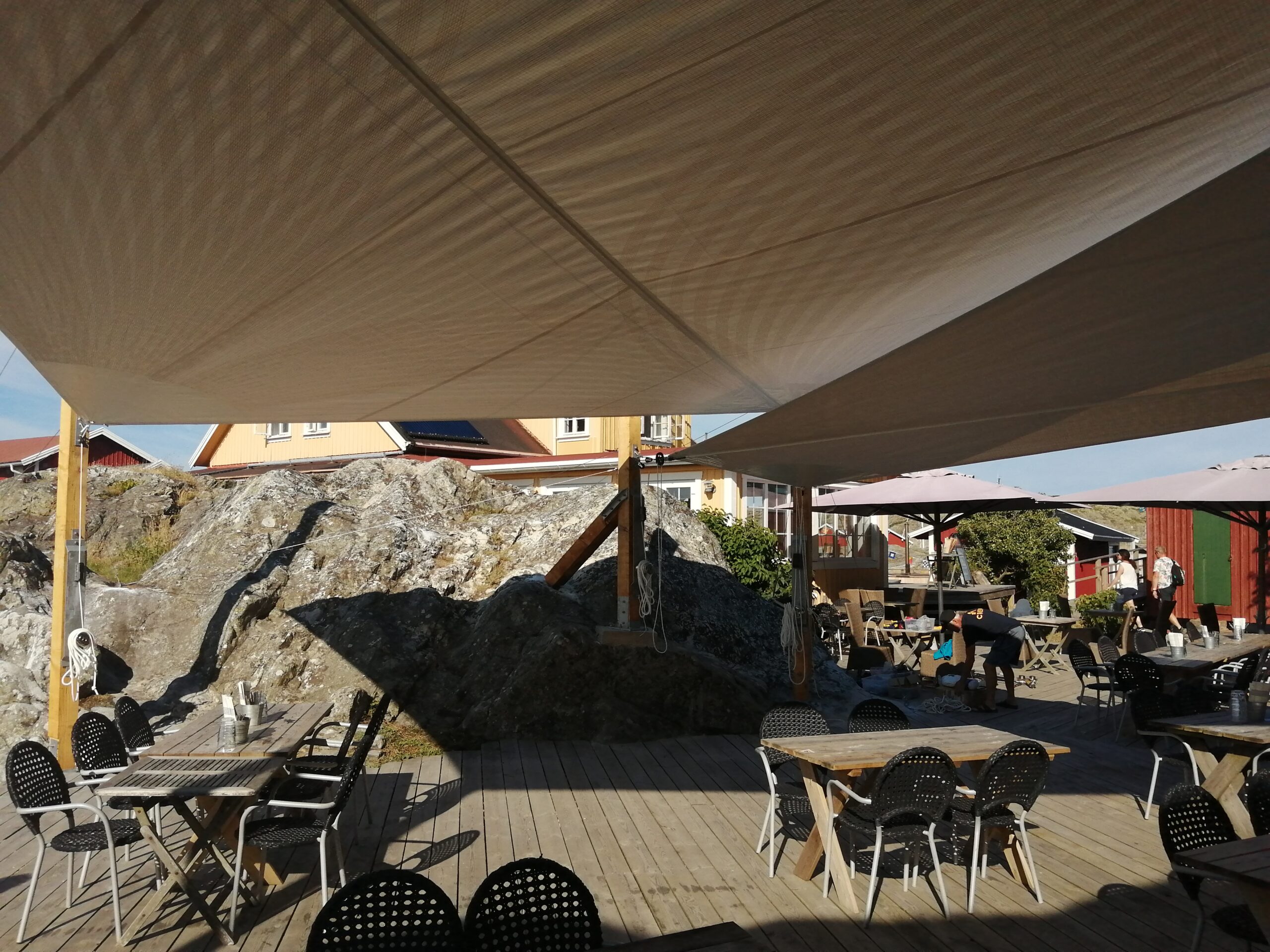 Utomhusrestaurang skyddad av ett stort beige solsegel med pergolamarkis, mot en bakgrund av klippor och trähus.